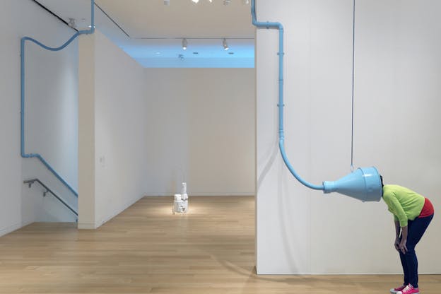 A figure hunches on the side of a white gallery room with their head inside a blue nozzle connected to a thin tube attached to the walls of the gallery, disappearing to the direction of a descending stairway on the left.