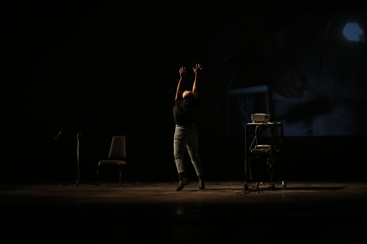 A performance still of Leslie Cuyjet in the middle of a darkened space, holding her arms in the air and looking up. Her body is angeled away from the camera and she wears a black t-shirt and green pants. A minimal amount of warm light illuminates her while a chair, microphone stand, and shelving unit are covered in shadows beside her. A dim projection of a hand covers the back wall. 