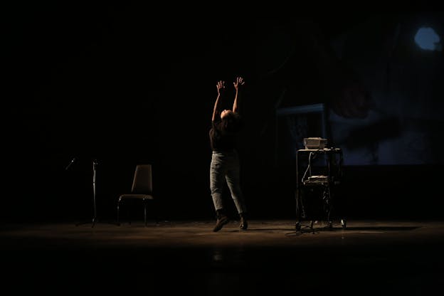 A performance still of Leslie Cuyjet in the middle of a darkened space, holding her arms in the air and looking up. Her body is angeled away from the camera and she wears a black t-shirt and green pants. A minimal amount of warm light illuminates her while a chair, microphone stand, and shelving unit are covered in shadows beside her. A dim projection of a hand covers the back wall. 