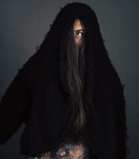 Portrait of Luciana Achugar in front of a slate grey background. She wears a fuzzy black garment which covers her arms, torso, head, and half of her face. One of her eyes is visible and open wide. Her long black hair covers the other eye. A brown and green marble textured garment is visible on her waist underneath the black fuzzy garment. 