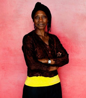 A portrait of Germaine Acogny looking into the camera with crossed arms. She wears a buttoned black sheer cardigan on top of a neon yellow tank top and her hair is wrapped in a bun. She stands against a pink rice paper background.