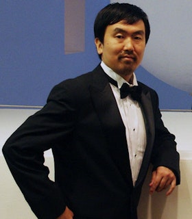 A portrait of Ei Arakawa against a white and blue wall. He is wearing a suit and a black bow tie and leans one elbow against the wall while keeping his other arm bent at his waist. 