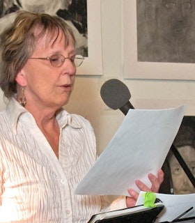 A side angle portrait of Rae Armantrout in front of a microphone, reading something off of a piece of paper. She has short blonde hair and wears dangling beaded earrings, wire rimmed glasses, and a textured white button down shirt. A few charcoal portraits hang on the wall behind her. 