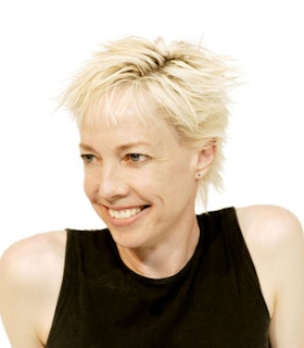 A portrait of Karole Armitage smiling to the left. She has a blonde pixie haircut and wears a black tank top.