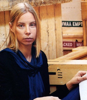 A portrait of Fia Backstrom in front of a wood plank wall and a wooden box on her right. She has medium length blonde hair and wears a black long sleeved shirt and a blue scarf. On her lap, she holds a piece of paper with illegible writing. 