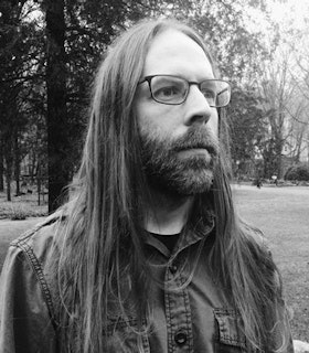 A black and white side angle portrait of Mick Barr in front of a tree lined grassy terrain. He has long straight hair and wears glasses and a button up shirt. 