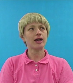 A portrait of Tamy Ben Tor with short blonde hair, wearing a pink polo shirt and standing in front of a bright blue background. She looks upwards and to the right, with her mouth held open, suggesting speech. 
