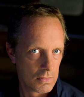 A close up portrait of Jonathan Bepler in front of a dark background. He wears a dark shirt and has short blonde hair and turqouise eyes. He looks intently at the camera with a thoughtful expression on his face.
