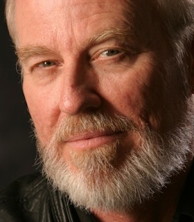 A close up portrait of John Bischoff in front of a dark background. He wears a black leather top and has short hair, blue eyes, and a beard. He looks directly at the camera and smiles. 