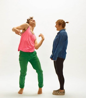 A performer in a pink top and green pants contorts their body upward while another performer in a blue sweater and black pants stands and looks toward them with a smile.