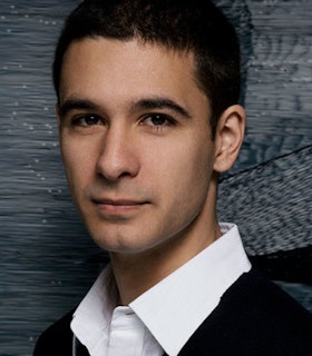 A portrait of Jonah Bokaer in front of a textured grey wall. He has short brown hair and wears a collared white shirt and dark sweater. 