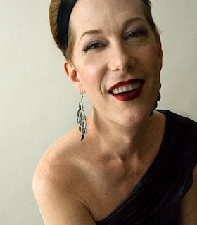 A portrait of Justin Vivian Bond against a cream wall wearing a black one shoulder top, dangling green earrings, red lipstick and a black headband. 