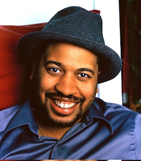 A portrait of Thomas Bradshaw against a white and red background. He has some facial hair and wears a grey fedora and blue silk shirt.