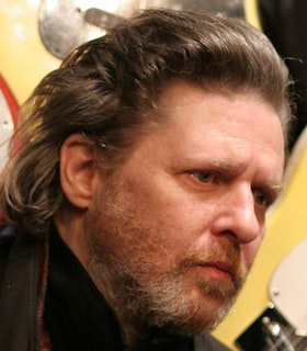 A side angle portrait of Glenn Branca in front of two light yellow electric guitars. He has medium length light brown hair and a close-cut beard. He wears a black shirt and a grey jacket. 