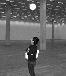 A black and white portrait of Paul Chan within an empty room with three columns. He crosses his arms over his chest and looks up towards a circle of light on the ceiling. He has short dark hair and wears a black sweater, dark jeans, and carries a white bag. 