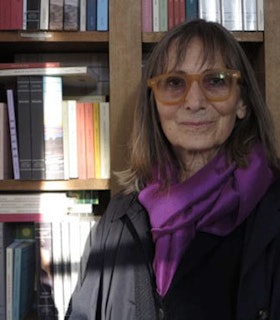 A portrait of Norma Cole in front of a filled bookshelf. She has shoulder length brown hair and wears orange glasses, a purple scarf, and a black jacket. 