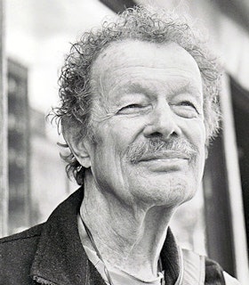 A black and white portrait of Jack Collom in front of a blurred hallway. He has short curly hair and a mustache and is wearing a collared jacket. 