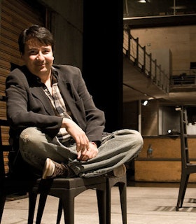 Martin Acosta sits cross legged on a black chair in a dimly lit space. Behind him, there is a staircase and another chair. He smiles warmly and wears converse sneakers, grey pants, a plaid shirt, and a grey blazer. He has short brown hair. 