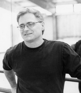 A black and white portrait of Terry Creach in front of a mirrored wall in a dance studio. He has short hair and wears a dark sweatshirt and thin rectangular wire rimmed glasses. He leans on a barre in the studio and tilts his head slightly downward and to the left, looking at something beyond the frame of the image and smiling slightly. 