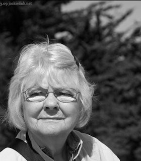 A black and white portrait of Dahlen Beverly in front of blurred foliage. She has short white hair and wears thin wire glasses, a black barrette and a white collared shirt. 