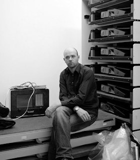 A black and white portrait of Rhodri Davies sitting in front of a shelf. He is bald and wears a black button up and dark jeans. He has his legs crossed at the knees and hist arms are crossed at the elbows and rest on his waist. 