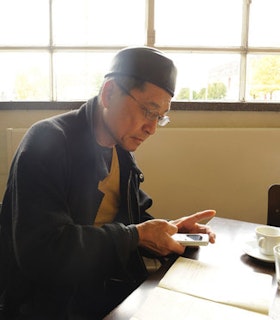 Portrait of David Diao looking down at a device, wearing a cap and a dark jacket. The artist sits at a table in front of a bright window.