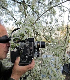 A side angle portrait of Nathaniel Dorsky holding a camera to his eye as he stands within tree branches filled with white flowers. There is a lake with a small boat behind the tree branches. 