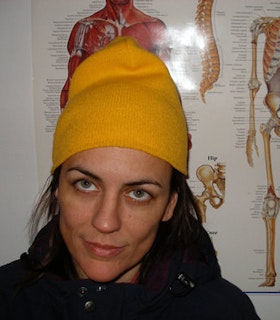 A portrait of DD Dovillier wearing a yellow beanie and a black parka, standing in front of an anatomy poster. She looks upwards and smiles.  