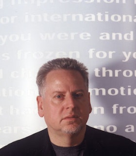 A portrait of Arnold Dreyblatt in front of a purple background with white text written across it. Dreyblatt wears a black shirt and has short gray hair. He looks directly at the camera.