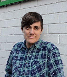 Portrait of Faye Driscoll in front of a white brick wall. She has short brown hair and wears a blue and turquoise flannel. 