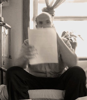 A black and white portrait of Kevin Drumm sitting on a chair in front of a window with a plant on the sill. He holds a blurred piece of paper up to his face, partially obscuring him. He is bald and wears a grey button up shirt and black pants. 