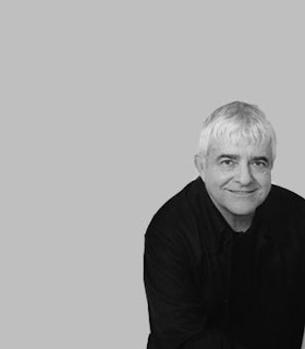 A black and white portrait of William Duckworth smiling in front of a grey background. He has white hair and wears a black top. 