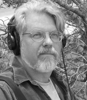 A close up, black and white portrait of David Dunn in front of a pine tree. He has short white hair and a gotee and wears headphones, thin wire glasses, a dark jacket and a white t-shirt. 