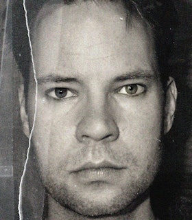 A close up black and white portrait of David Dupuis against a bare dark background. Dupuis looks directly at the camera with an intent expression on his face. There is a rip down the entirety of the photograph on the left side.