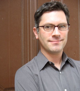 A portrait of founder John Collins in front of a brown background. He has short grey hair and wears thin brown glasses and a grey button up shirt. 