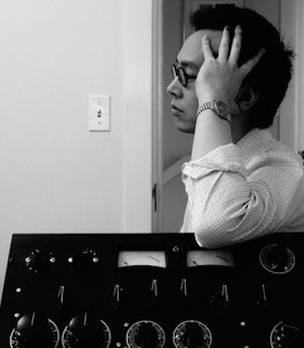 A black and white profile of James Fei resting their head in their hand, wearing a rolled up button up shirt. Their arm is resting on sound equipment.