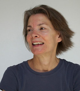 A close up portrait of Molissa Fenley wearing a blue shirt in front of a light grey background. She has short brown hair and smiles and she turns slightly away from the camera and looks at something beyond the frame of the image. 