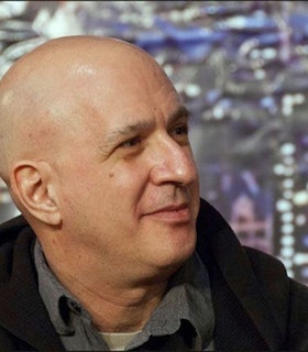 A three quarter profile portrait of David First against a metallic silver background. He is bald and wears a grey collared shirt and a dark sweatshirt. He smiles slightly and looks at something beyond the frame of the photograph.