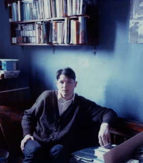 A portrait of Luke Fowler sitting in front of a blue wall with books shelves above him to the left. He has short brown hair and wears a brown buttoned cardigan, jeans, and a white button up shirt. He sits on a red velvet patterned seat with even more books on it. 
