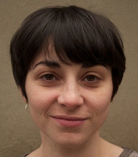 A close up portrait of Beth Gill in front of a light brown wall. She has short brown hair and wears small earrings. 