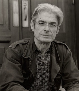 A black and white portrait of John Godfrey in front of two doors. He has short white hair which is parted in the middle and wears frame-less glasses, a white and black patterned button up shirt, and a jacket with epaulettes.
