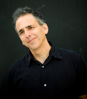 Michael Gordon stands in front of a black background. He has short grey and black hair and wears a black collared shirt. He smiles slightly and tilts his head to the left.