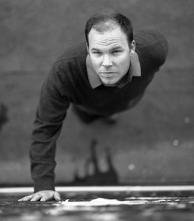 A black and white portrait of David Grubbs standing with on hand against the wall, looking up at the camera. He has short hair and wears a sweater and collared shirt. 