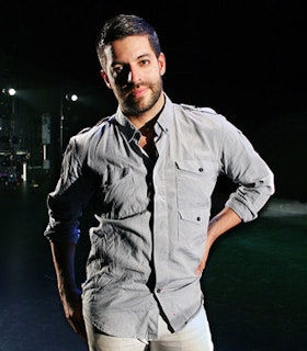 A portrait of Miguel Gutierrez on a dark, empty stage lit only by two spotlights. He has short black hear, wears a grey button up and grey pants, and is resting one hand on his hip.  