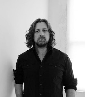 A black and white portrait of Wade Guyton standing against a light background. He has curly shoulder-length hair and wears a buttoned black shirt.