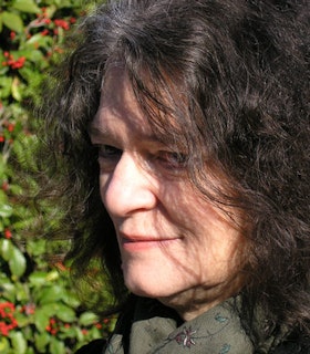 A close-up side portrait of Carla Harryman against a fruit tree background. She smiles and looks at a 45-degree angle.