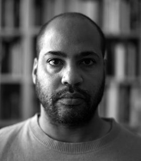 Close up black and white portrait of David Hartt in front of blurred bookshelves. The artist has a close cut beard and wears a plain T-shirt.
