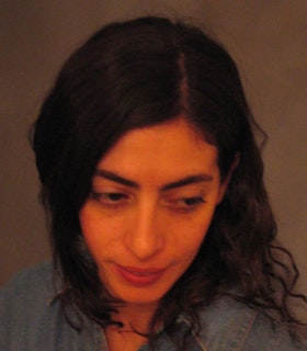 A high angle orange hued portrait of Maria Hassabi. She looks down to the right. She has medium length curly black hair and wears a denim shirt.  