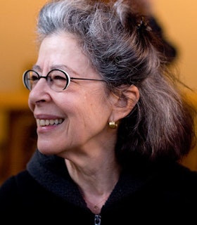 A side angled portrait of Deborah Hay in front of a blurred orange background. She has grey hair that is partially pulled into a top knot, thin black oval glasses, small gold hoop earrings and wears a zippered black sweater. 