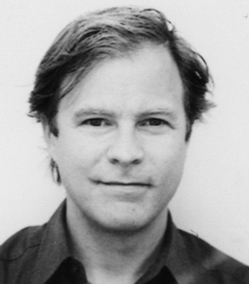 A close up black and white portrait of David Henderson smiling and looking directly at the camera against a white wall. He wears a dark collared shirt and has short hair.
